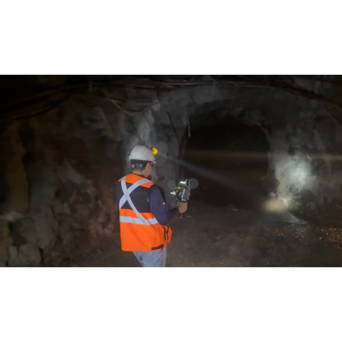 A person in a white hard hat and high-visibility orange safety vest stands inside a dark Quarrying tunnel holding a FJD Trion T1 laser scanning that emits a bright beam of light.