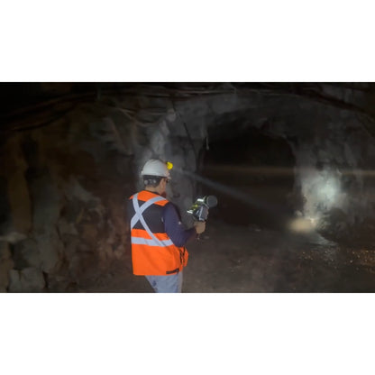 A person in a white hard hat and high-visibility orange safety vest stands inside a dark Quarrying tunnel holding a FJD Trion T1 laser scanning that emits a bright beam of light.