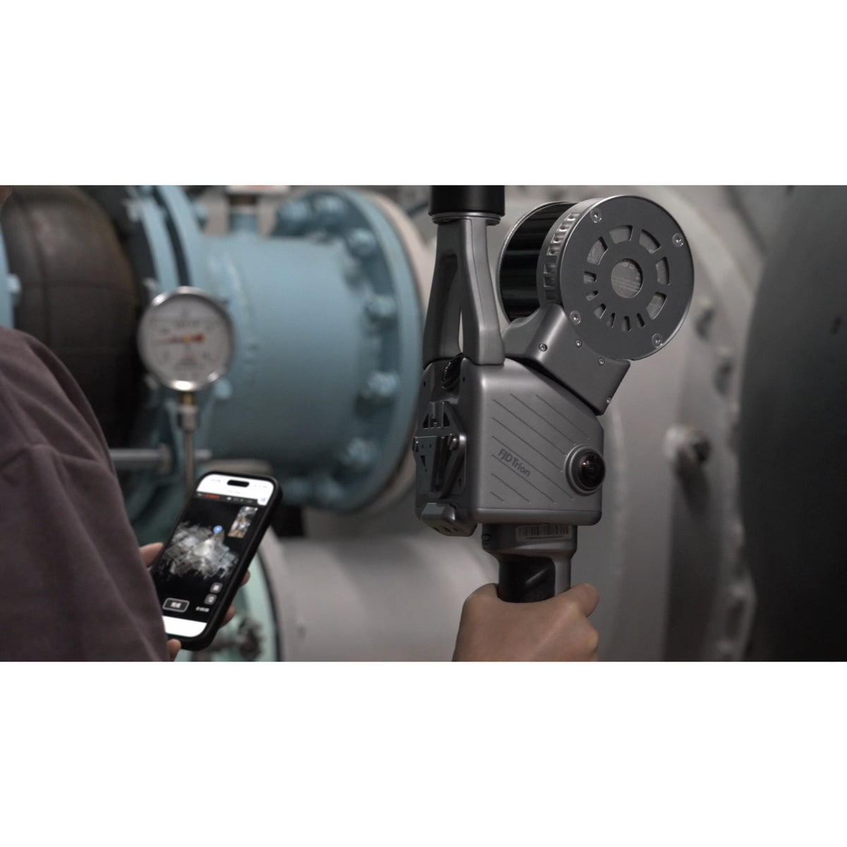A person holding a FJD Trion T1 hybrid LiDAR scanner in handheld configuration and using a smartphone in one hand for checking with the FJD Trion App, photographed in an industrial setting with large metal machinery in the background.