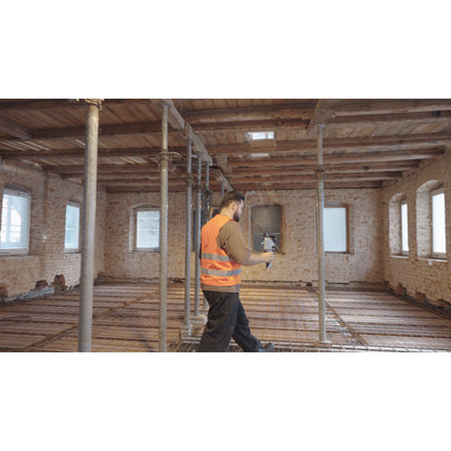 A construction worker wearing a high-visibility orange safety vest walks through a large, unfinished brick room with Operator using the FJD Trion X1 to capture an unfinished building interior for as-built documentation